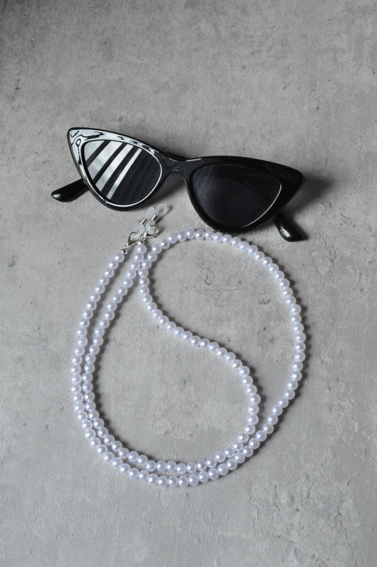 On a grey background, a pair of black cat-eye glasses is displayed with the Faux Pearl Glasses Holder draped over them. The chain features elegant pearls and a sophisticated design.