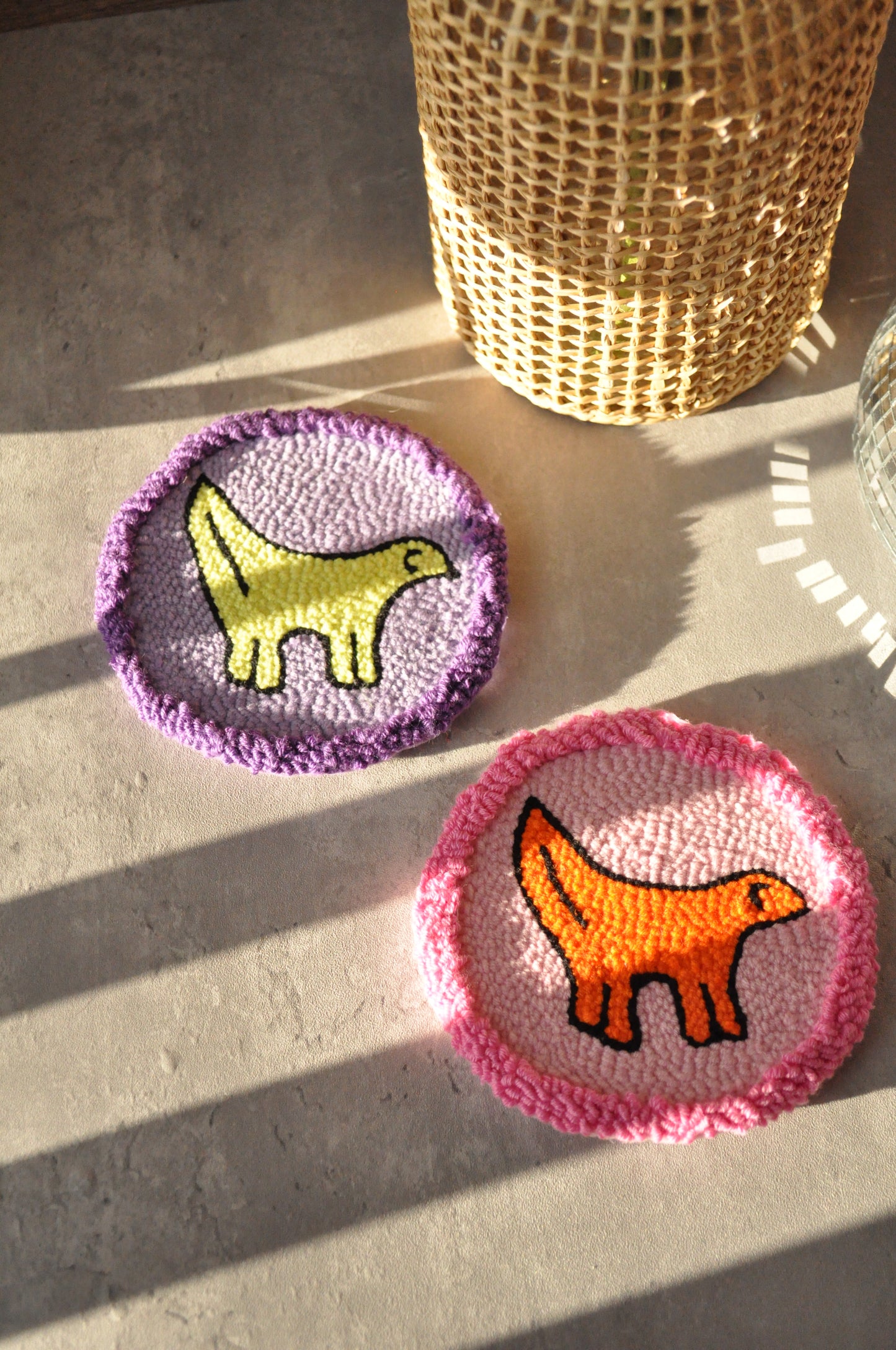 Two handmade punch needle coasters featuring a Lambanana design are placed on a textured grey surface. One coaster has a yellow Lambanana on a purple background, while the other has an orange Lambanana on a pink background. A woven rattan-wrapped glass vase with greenery is in the background, and a small disco ball reflects light on the right side of the image. The setting has a warm, natural aesthetic with soft lighting.