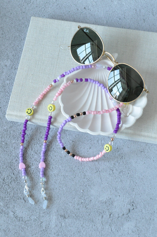 On a grey background, a textured notebook and a shell trinket dish are positioned. Resting on top is a pair of round sunglasses adorned with a lilac beaded glasses chain featuring smiley beads and rose gold accents.