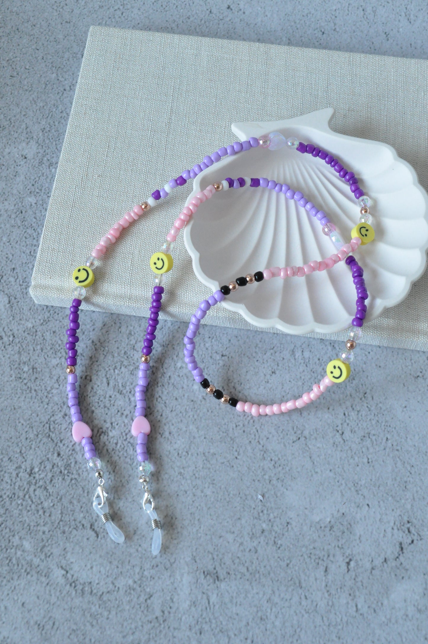 On a grey background, a textured notebook and a shell trinket dish are positioned. A lilac beaded glasses chain featuring smiley beads and rose gold accents is draped over the items.