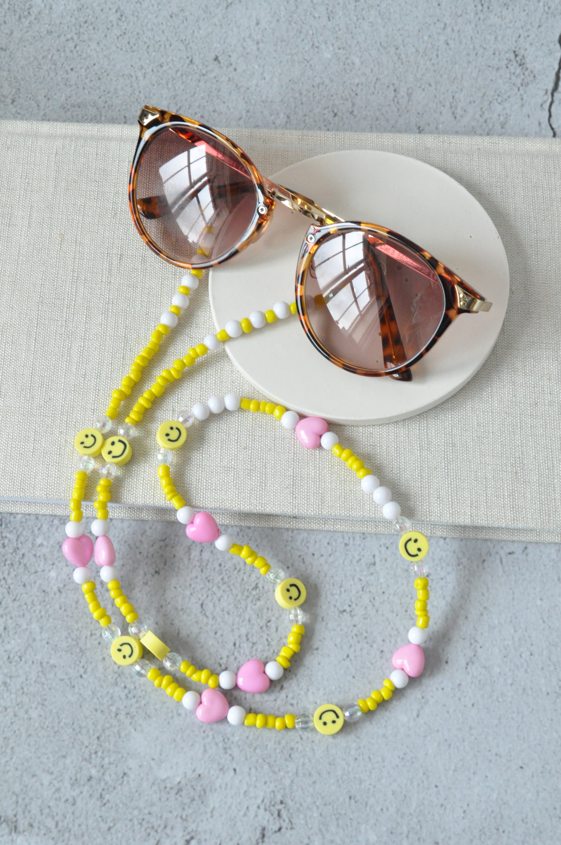 On a grey background, a textured notebook is positioned with a pair of brown sunglasses and the smiley beaded glasses chain displayed on top. The chain features yellow seed beads, smiley faces, and plated connectors.