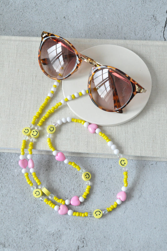 On a grey background, a textured notebook is positioned with a pair of brown sunglasses and the smiley beaded glasses chain displayed on top. The chain features yellow seed beads, smiley faces, and plated connectors.