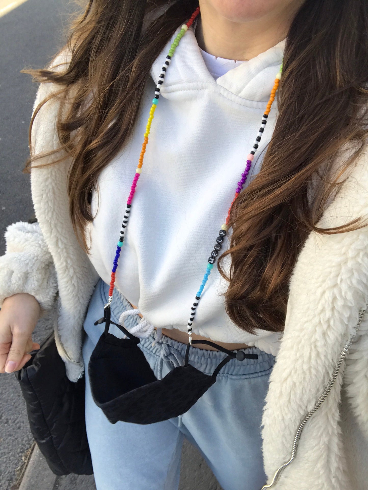 A long-haired girl models the beaded glasses chain, personalised with the name "Melly," as a mask necklace against a street background. The vibrant beads are draped stylishly around her neck, highlighting its unique and colourful design.