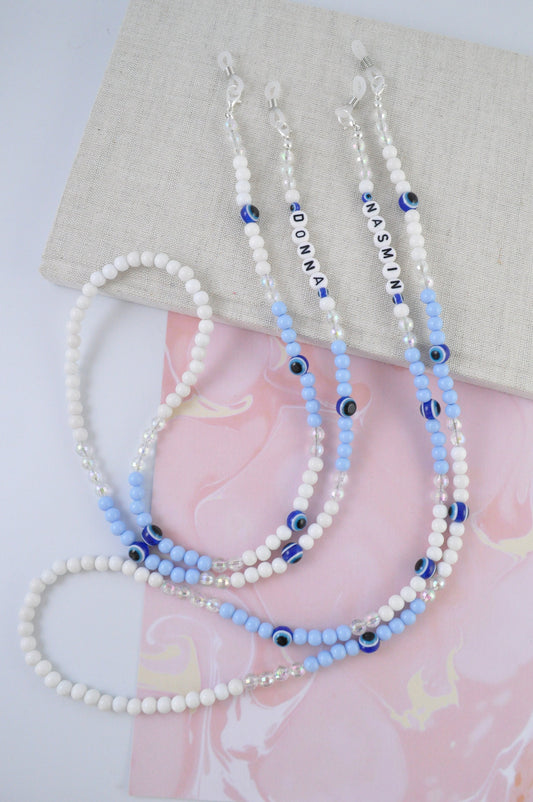 On a white and pink background, a textured notebook is positioned. Resting on top are two personalised Evil Eye Beaded Glasses Chains, each featuring different names, high-quality blue beads, and the iconic Evil Eye symbol.