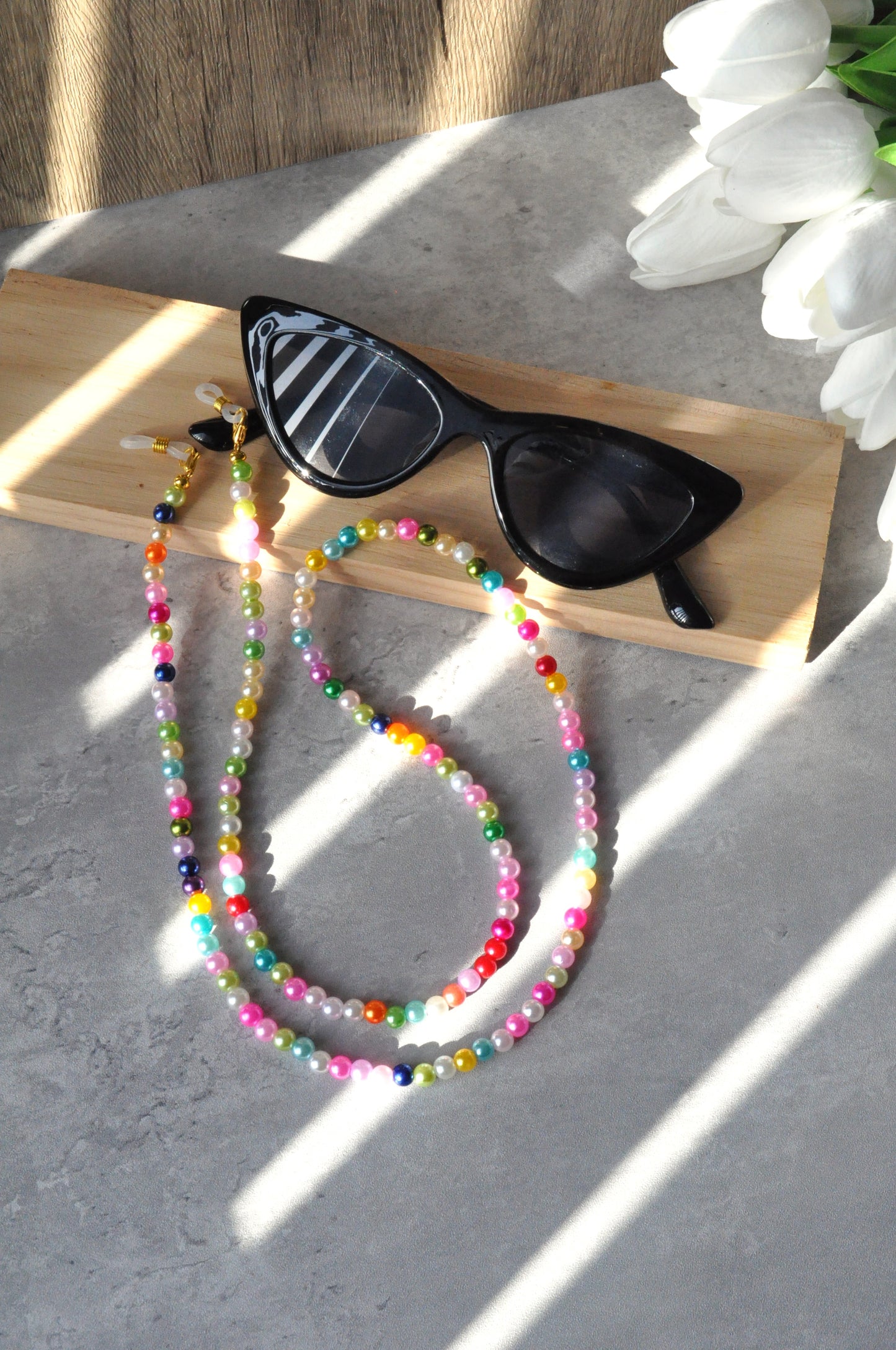 Black sunglasses with colorful beaded chain on a wooden block with white tulips in the background