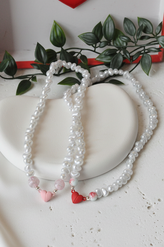 Two pearl necklaces with heart decorations on a white surface with green leaves and a red heart in the background.