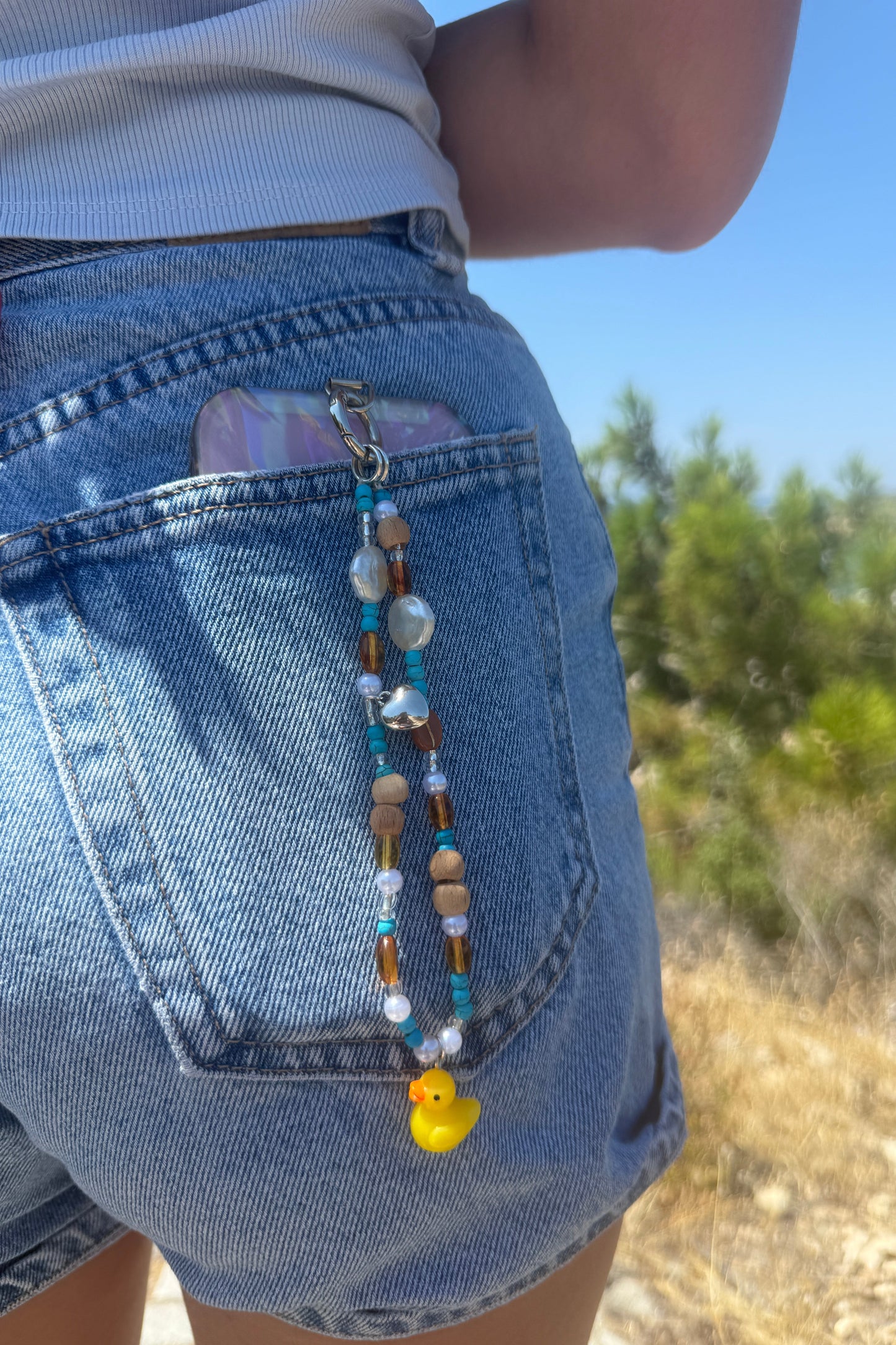 A close-up of a person wearing blue denim shorts outdoors, with a beaded phone strap or lanyard draped across the back pocket. The strap features brown, white, and blue beads, a small clear stone, and a small yellow duck charm.