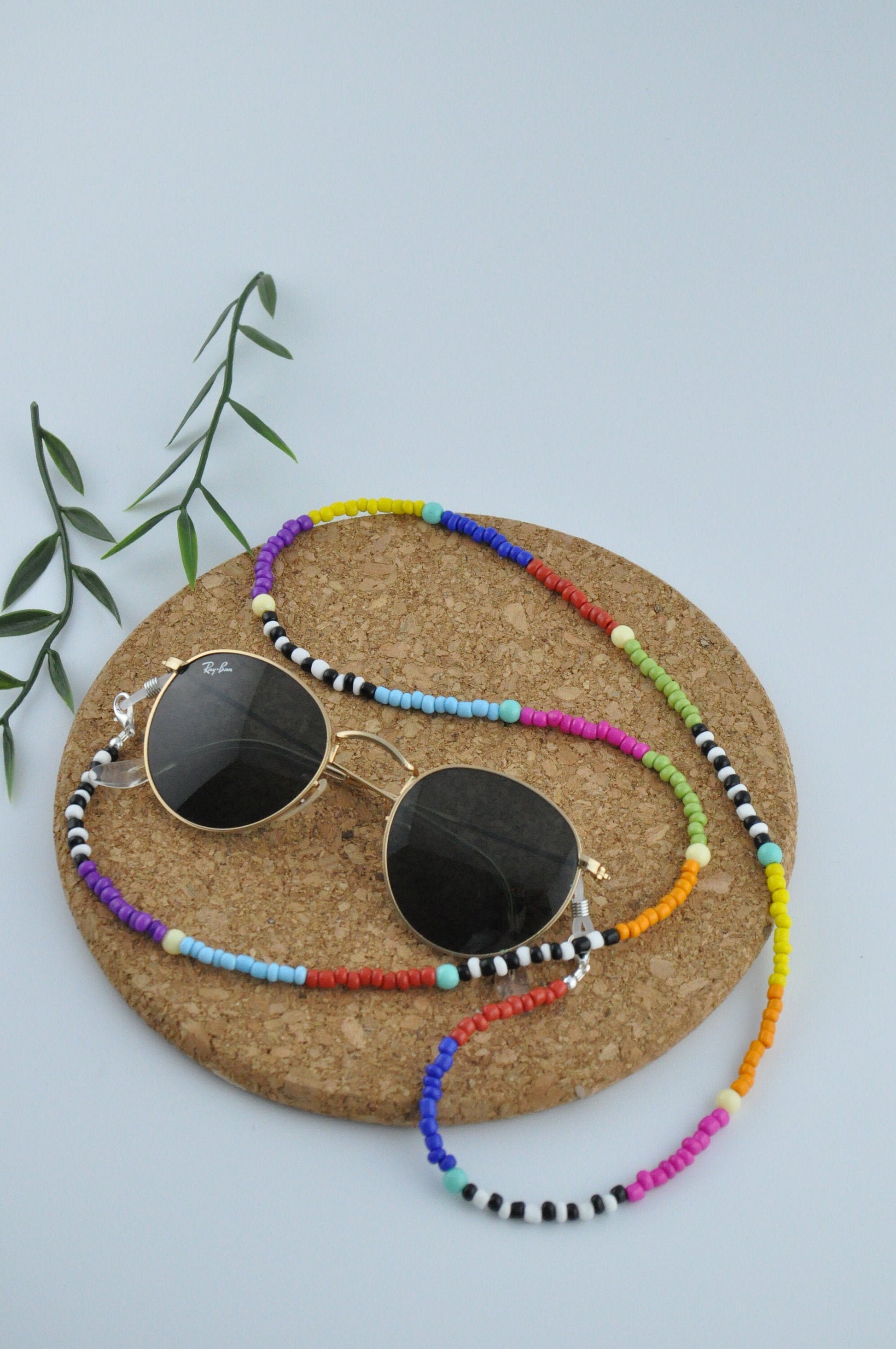 On a white background, a round cork is positioned with a pair of black glasses featuring gold detailing resting on top. The glasses are adorned with a vibrant beaded chain, adding a splash of colour to the elegant design.