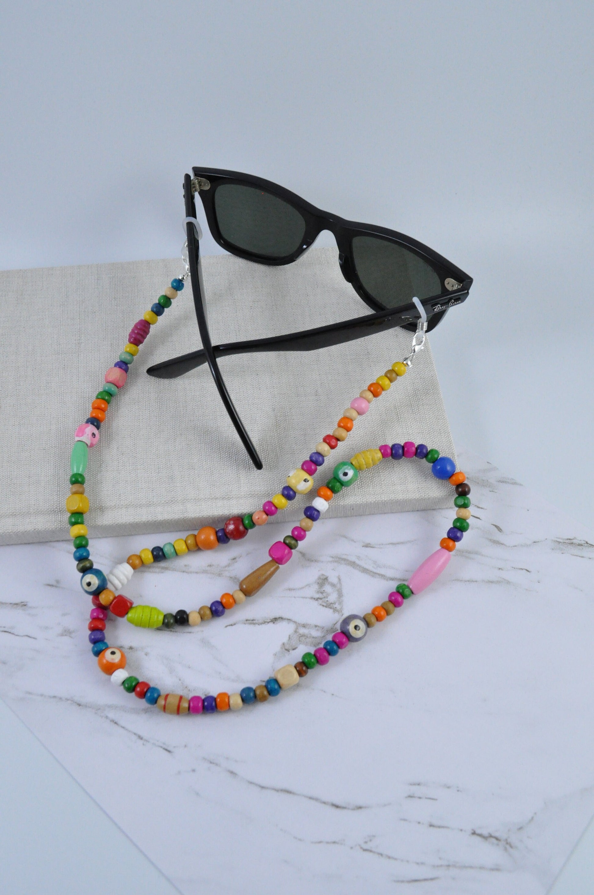 On a grey marble background, a textured notebook is positioned with a pair of black sunglasses and the Wooden Beaded Sunglasses Chain displayed on top. The chain features high-quality wooden beads and complements the sunglasses.